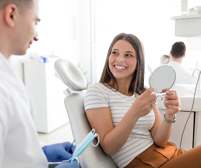 Young woman looking at a mirror and loving her new smile after recent cosmetic dentistry while in dentist chair.